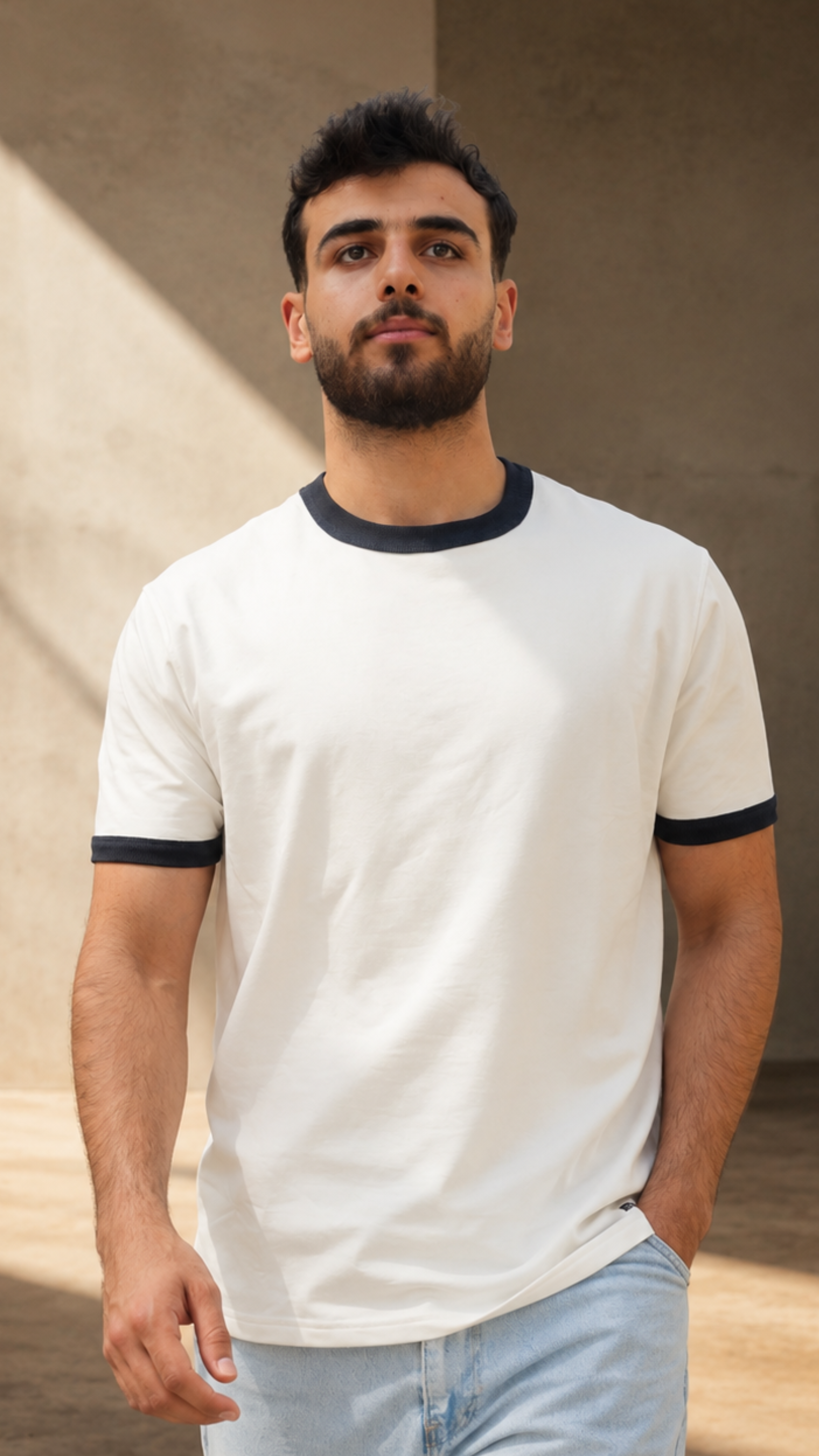 A man wearing a white ringer t-shirt with dark navy trim on the collar and sleeve cuffs, paired with light blue jeans, standing in a sunlit concrete setting.