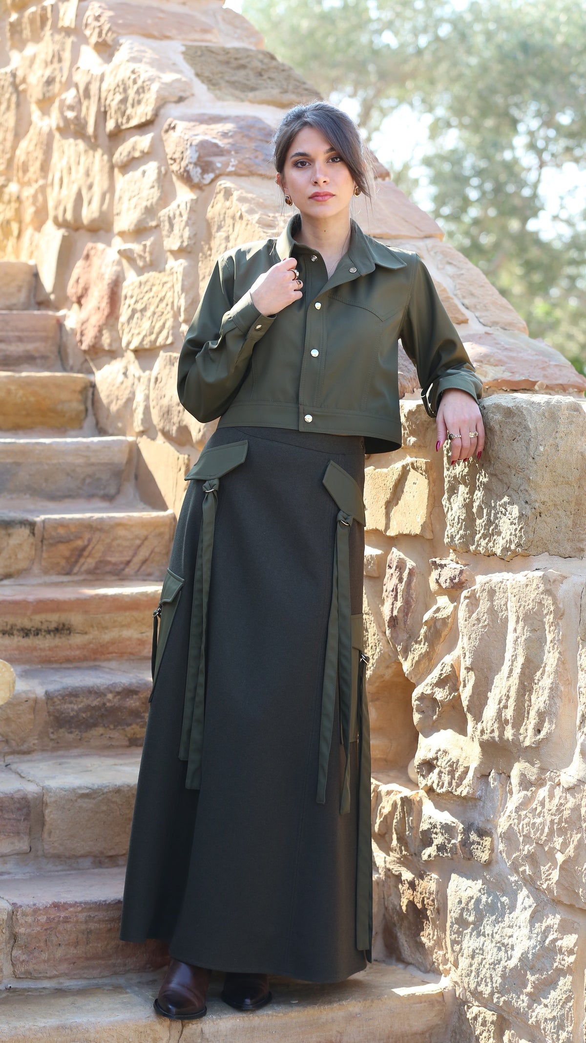 A woman stands on stone stairs wearing an olive green cropped button-up shirt and a matching dark olive green maxi skirt with side tie details.