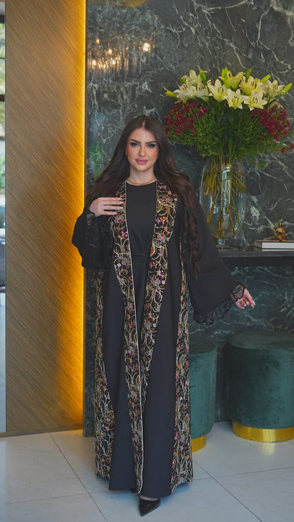 A woman models a long black abaya with a beige and brown patterned outer layer draped over her shoulders, standing in an elegant lobby with a marble wall, floral arrangement, and green velvet stools.