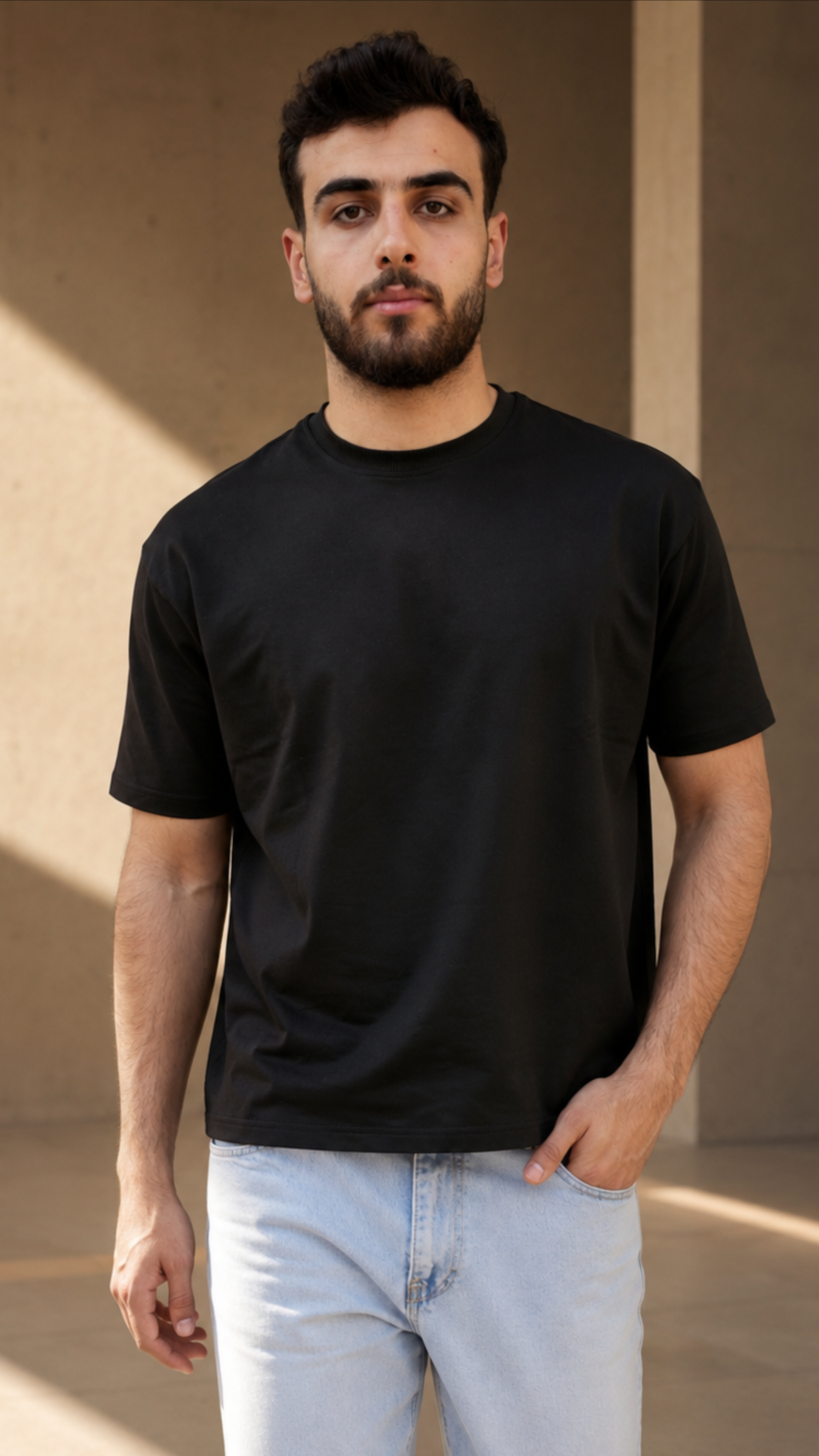 A man wearing a plain black short-sleeve crewneck t-shirt and light blue jeans, standing in a modern concrete structure with warm sunlight and shadows.