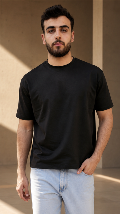 A man wearing a plain black short-sleeve crewneck t-shirt and light blue jeans, standing in a modern concrete structure with warm sunlight and shadows.
