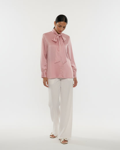 Woman wearing a pink blouse with a bow tie and white pants on a white background