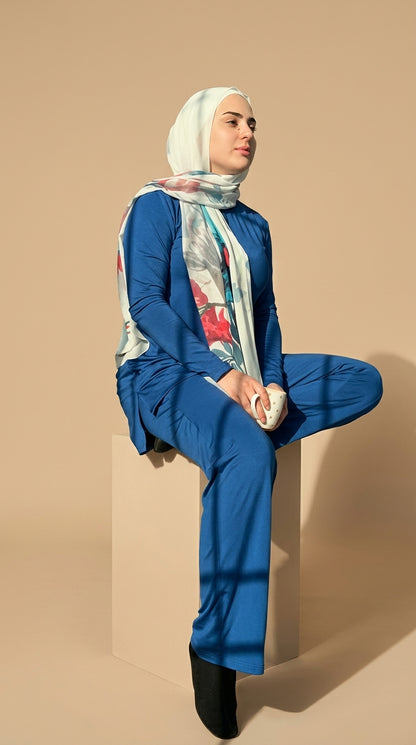 Woman wearing a blue outfit and patterned hijab sitting against a beige wall.