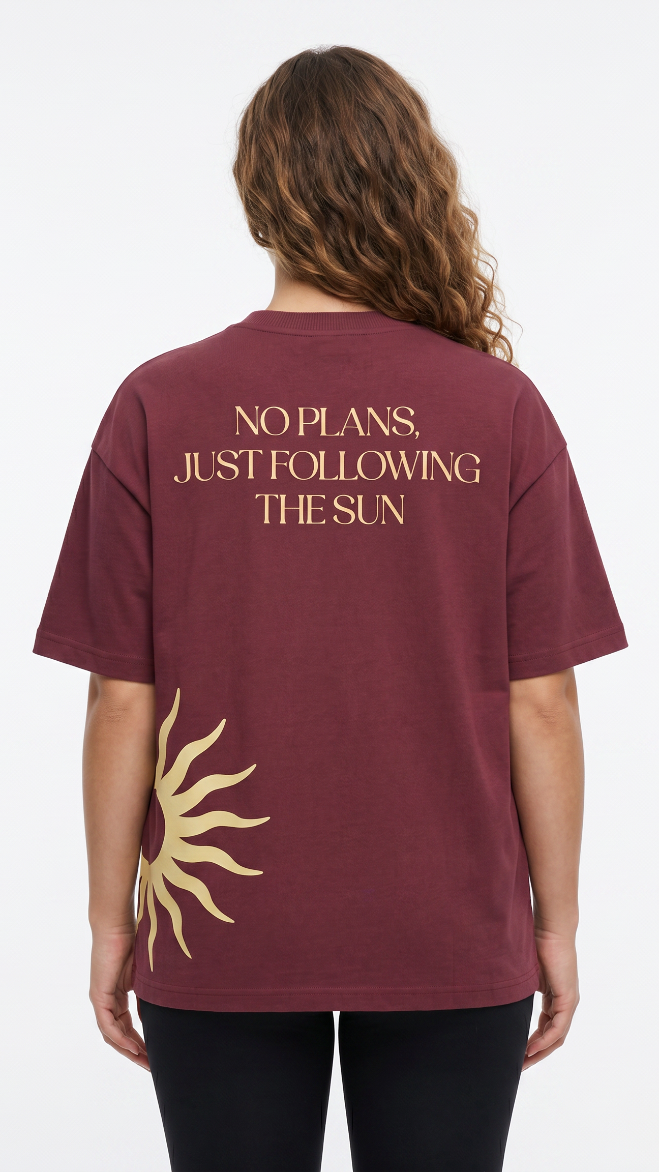 A woman viewed from the back wearing a maroon short-sleeve T-shirt with gold text reading 'NO PLANS. JUST FOLLOWING THE SUN' and a gold sun graphic near the lower back, paired with black leggings against a light gray studio background.
