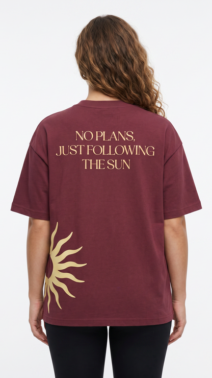 A woman viewed from the back wearing a maroon short-sleeve T-shirt with gold text reading 'NO PLANS. JUST FOLLOWING THE SUN' and a gold sun graphic near the lower back, paired with black leggings against a light gray studio background.