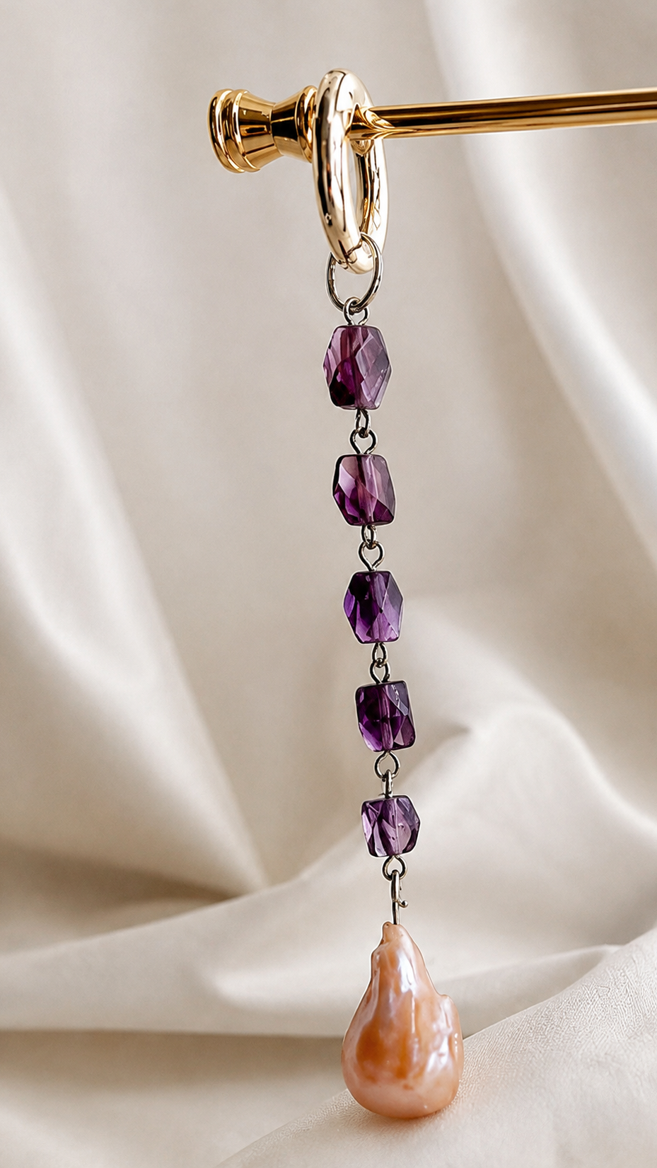 A long drop charm with five faceted purple beads connected by dark metal links and a large teardrop-shaped peach-pink pearl at the bottom, hanging from a gold curtain rod against draped cream fabric.