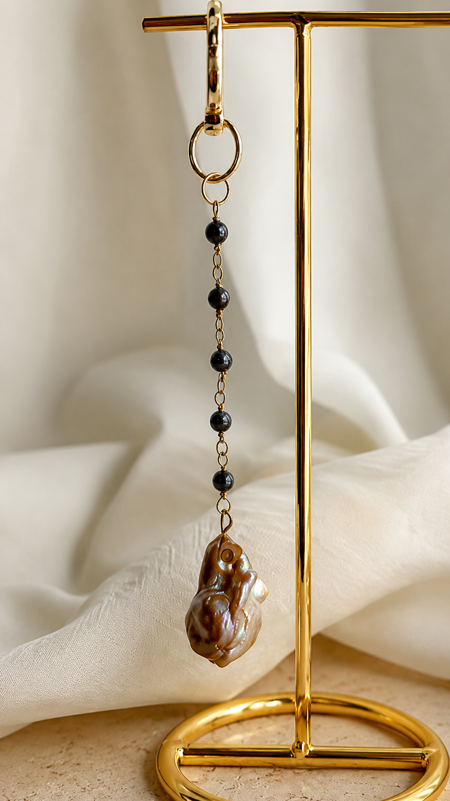 A long dangle earring with a gold-tone clasp, small dark beads, and a chunky brown agate pendant, displayed on a gold stand against a draped cream fabric background.