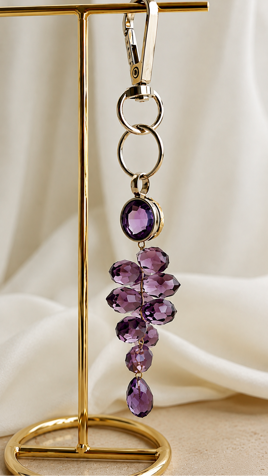A gold-tone keychain with a cluster of faceted purple amethyst gemstones, hanging from a gold stand against a draped cream fabric background.