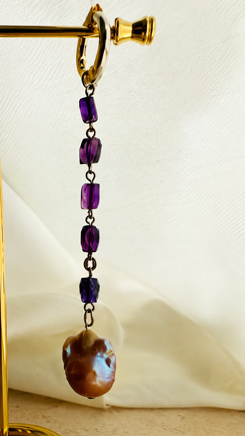 A single dangling earring displayed on a gold stand against a draped cream fabric background. The earring has a gold hoop at the top, several purple faceted beads, and a large irregular brown shell-like charm with iridescent blue highlights.