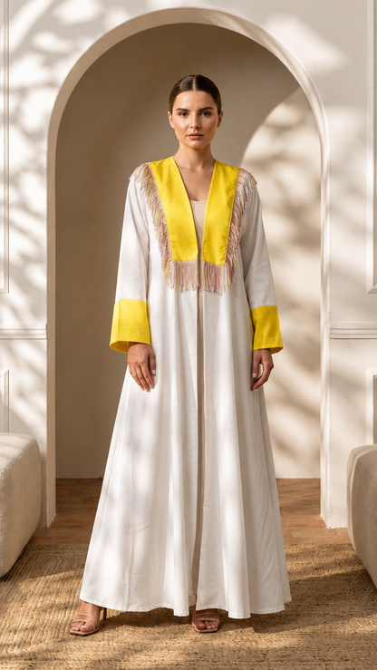 A woman stands in a real interior setting with an arched doorway, wall paneling, floor rug, and upholstered seating, wearing a long white caftan-style dress with bright yellow trim and embroidered details at the neckline and cuffs.