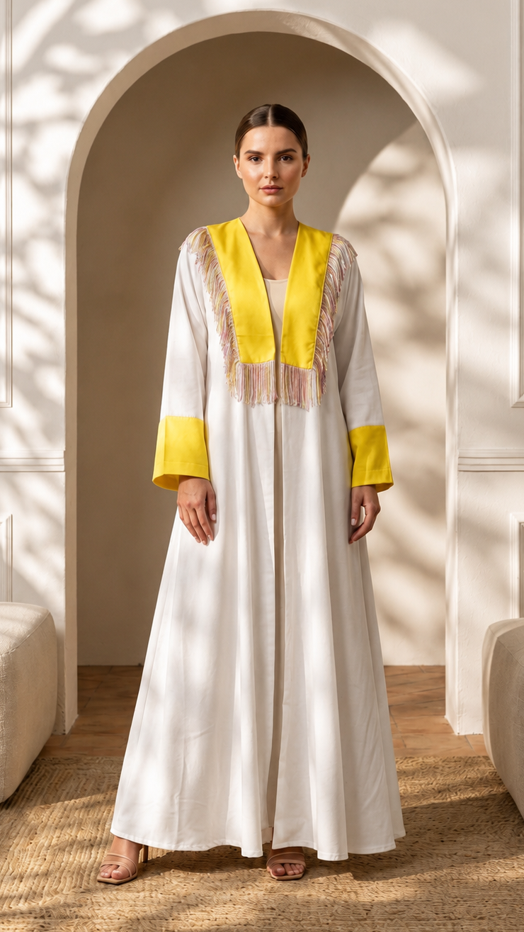 A woman stands in a real interior setting with an arched doorway, wall paneling, floor rug, and upholstered seating, wearing a long white caftan-style dress with bright yellow trim and embroidered details at the neckline and cuffs.