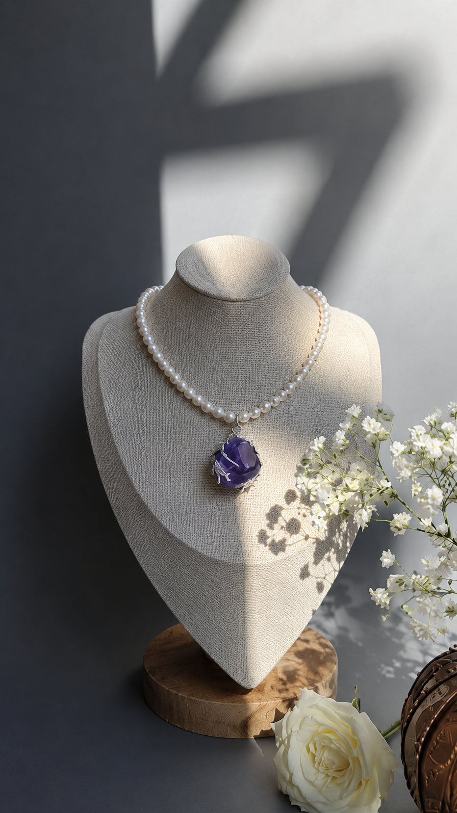 A pearl necklace with a large purple gemstone pendant displayed on a beige jewelry bust. The bust sits on a wooden slice stand, with white flowers and a brown textured object in the foreground against a dark gray background with soft shadows.