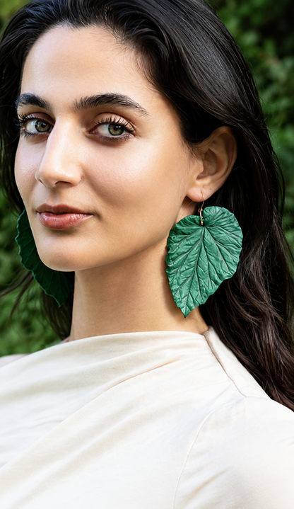 A close-up of a woman wearing large, textured green leaf-shaped drop earrings, with a soft-focus green outdoor background.