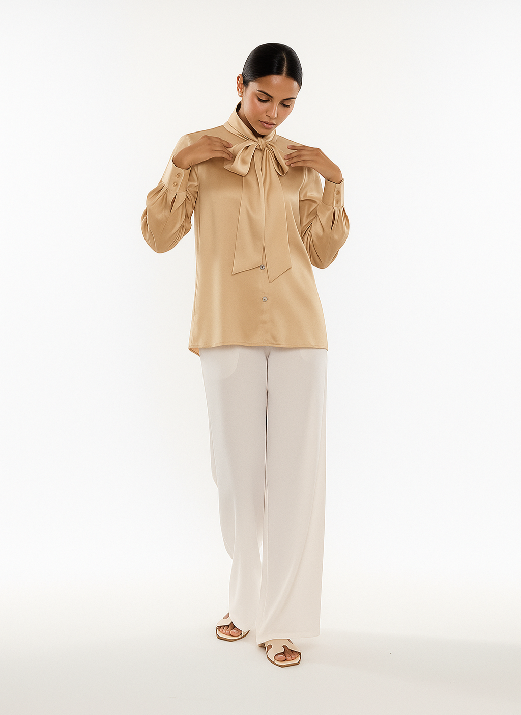 Woman wearing a beige blouse with a bow tie and white pants on a white background