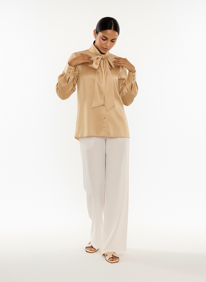 Woman wearing a beige blouse with a bow tie and white pants on a white background