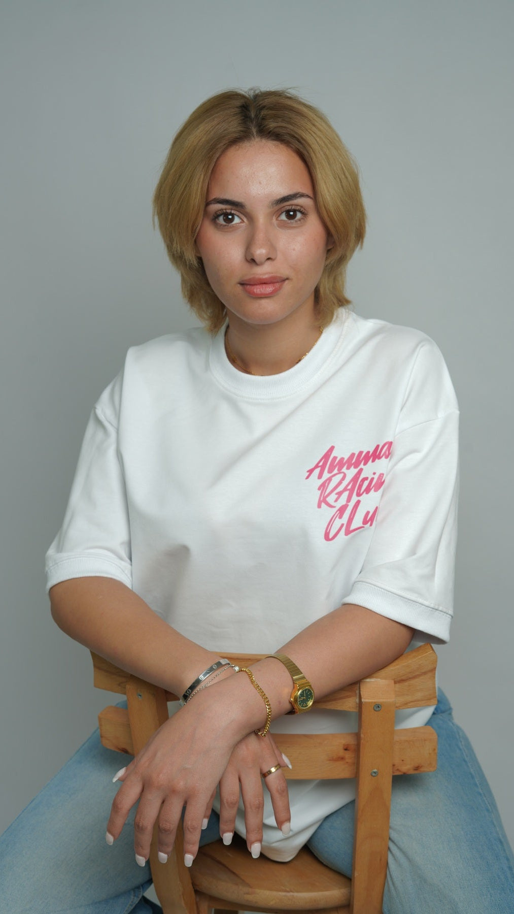 Person wearing a white t-shirt with pink text, sitting on a wooden chair against a gray background