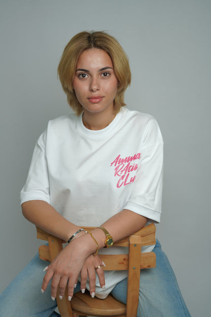 Person wearing a white t-shirt with pink text, sitting on a wooden chair against a gray background