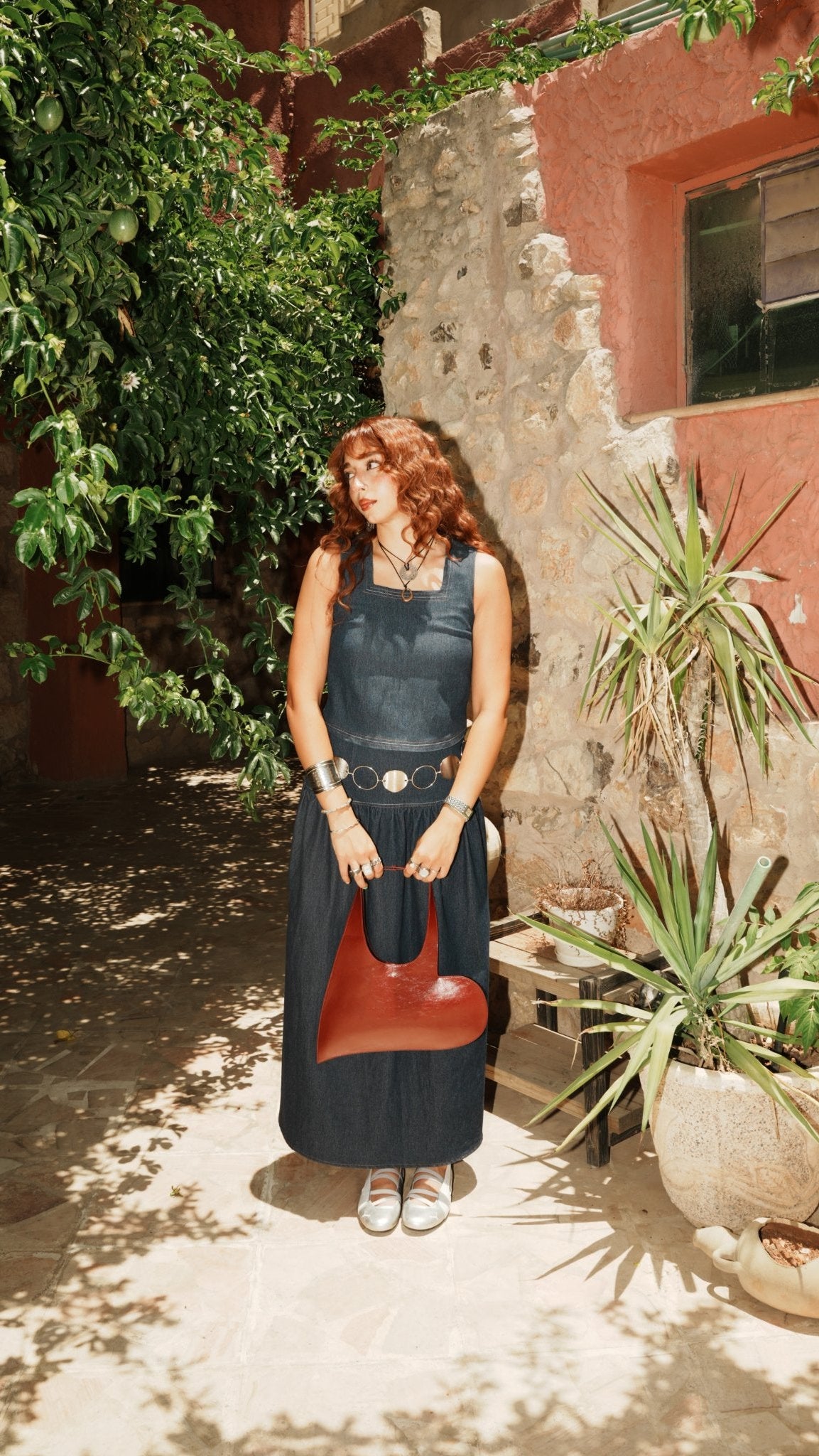 A woman with red hair stands in a sunlit courtyard wearing a dark navy sleeveless dress and holding a reddish-brown heart-shaped handbag. The background features a rustic terracotta wall, stone masonry, and potted plants.
