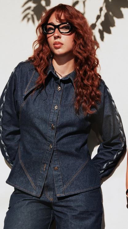 A woman with long wavy red hair and black glasses wearing a dark blue denim jacket with sheer black sleeves and matching dark blue jeans, standing against a white wall with leaf shadows.