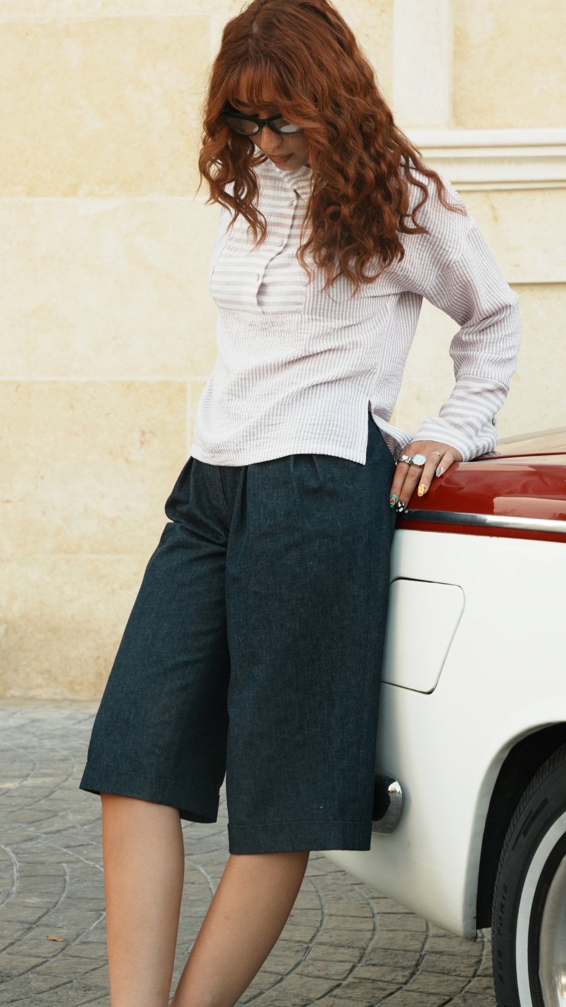 A woman with long wavy red hair and sunglasses leans against a vintage white car with a red stripe, wearing a white ribbed long-sleeve top and dark charcoal gray knee-length shorts, set against a beige stone wall.