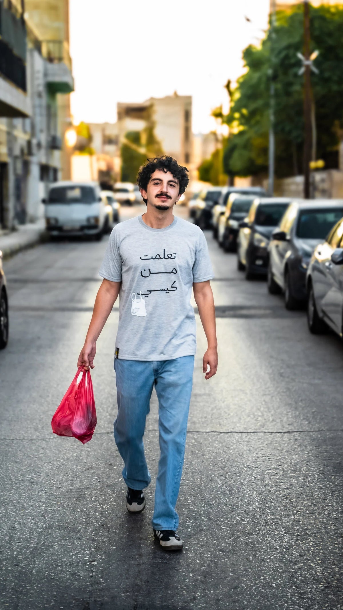 Man walking on a street holding a red bag, wearing a gray t-shirt with Arabic text.