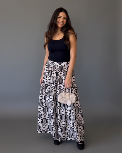 Woman wearing a black top and patterned skirt holding a clutch against a gray background