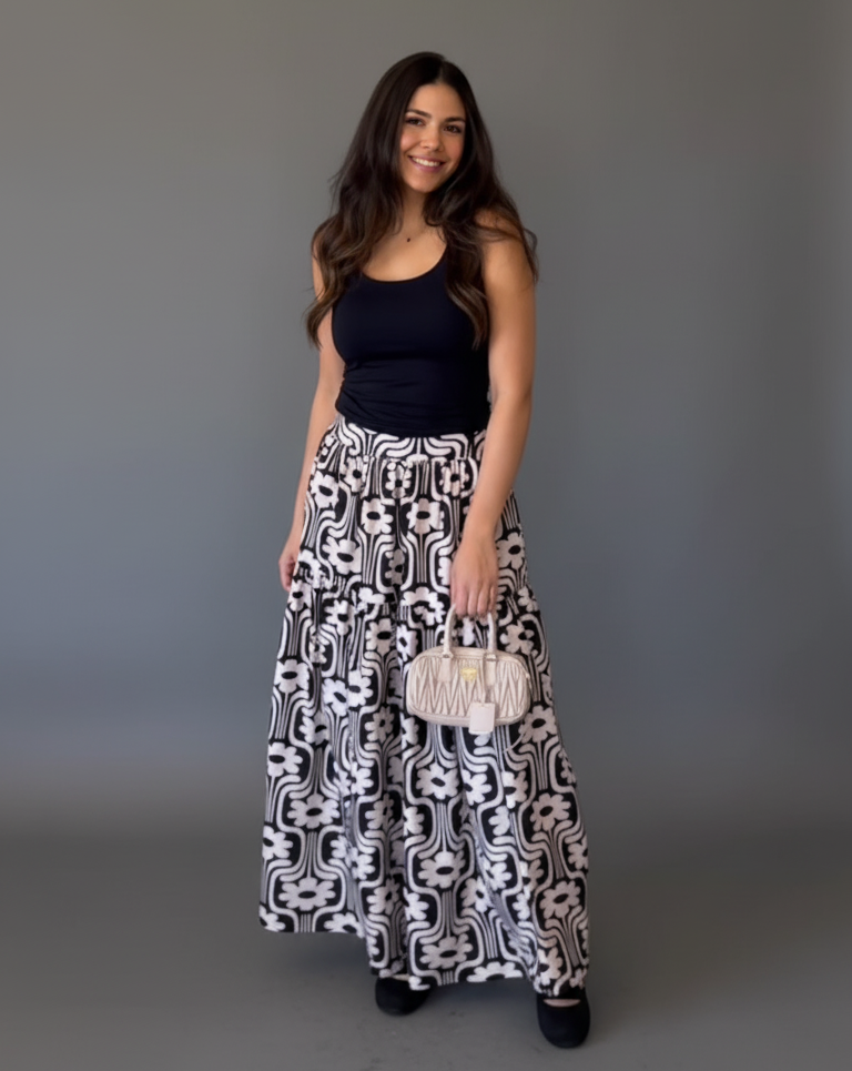 Woman wearing a black top and patterned skirt holding a clutch against a gray background