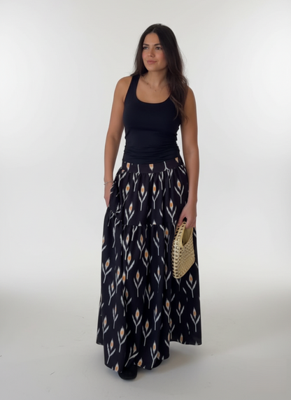 Woman wearing a black tank top and patterned long skirt holding a straw bag on a white background