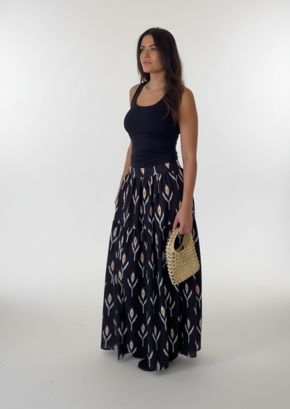 Woman wearing a black sleeveless top and patterned long skirt holding a straw bag on a white background