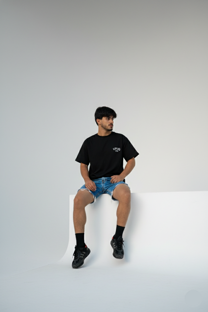Person wearing a black t-shirt and denim shorts sitting on a white surface with a plain background