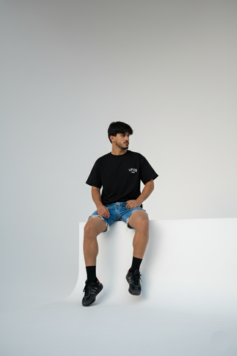 Person wearing a black t-shirt and denim shorts sitting on a white surface with a plain background