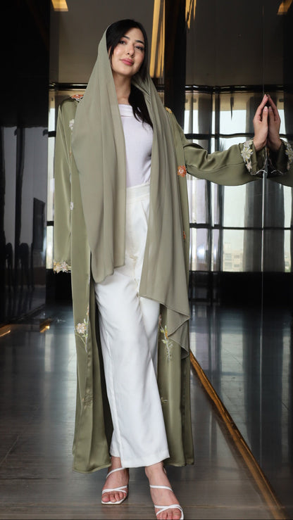 A woman wearing a long olive green open-front coat over a white outfit and white heeled sandals, standing in a dark indoor setting with reflective flooring.