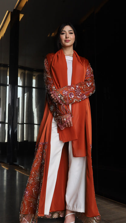 A woman models a traditional outfit featuring a rust-orange embroidered long coat over a white kurta and wide-leg trousers, set against a dark, modern interior background.