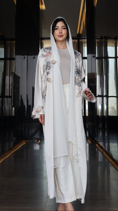 A woman wearing a long white traditional-style dress with floral embroidery and a matching head covering, standing in a dark interior hallway.
