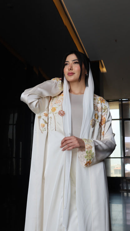 A woman wearing a white traditional outfit with floral embroidery and a matching sheer scarf, photographed indoors against a dark background.
