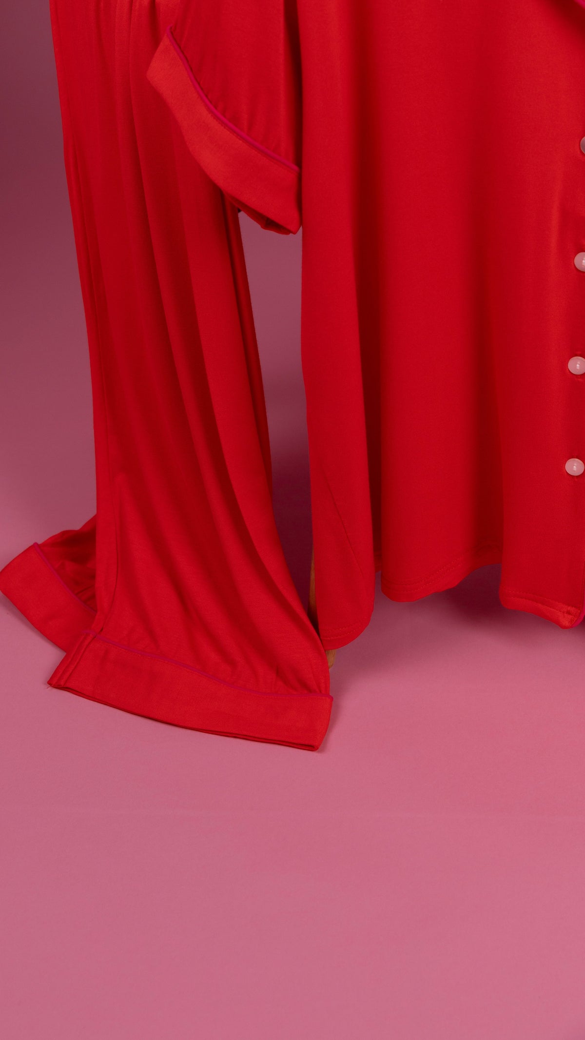 A close-up of a bright red flowing fabric garment or drape with a buttoned side detail, set against a solid pink studio background.