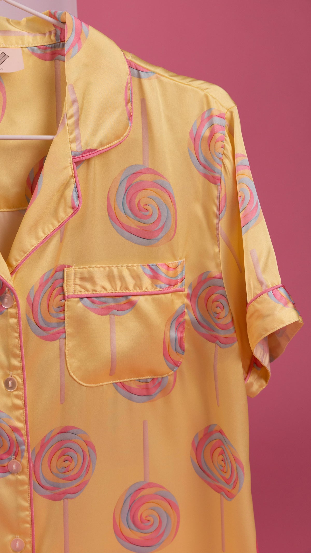A yellow satin pajama shirt with a pink and blue lollipop print, featuring a collar, button front, and chest pocket, shown hanging against a solid pink background.