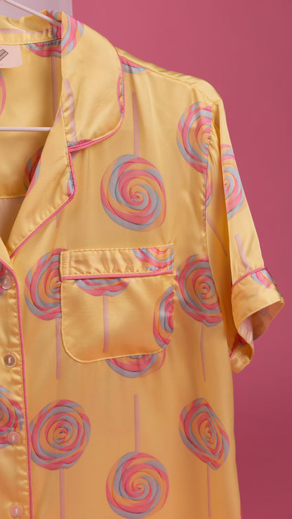 A yellow satin pajama shirt with a pink and blue lollipop print, featuring a collar, button front, and chest pocket, shown hanging against a solid pink background.