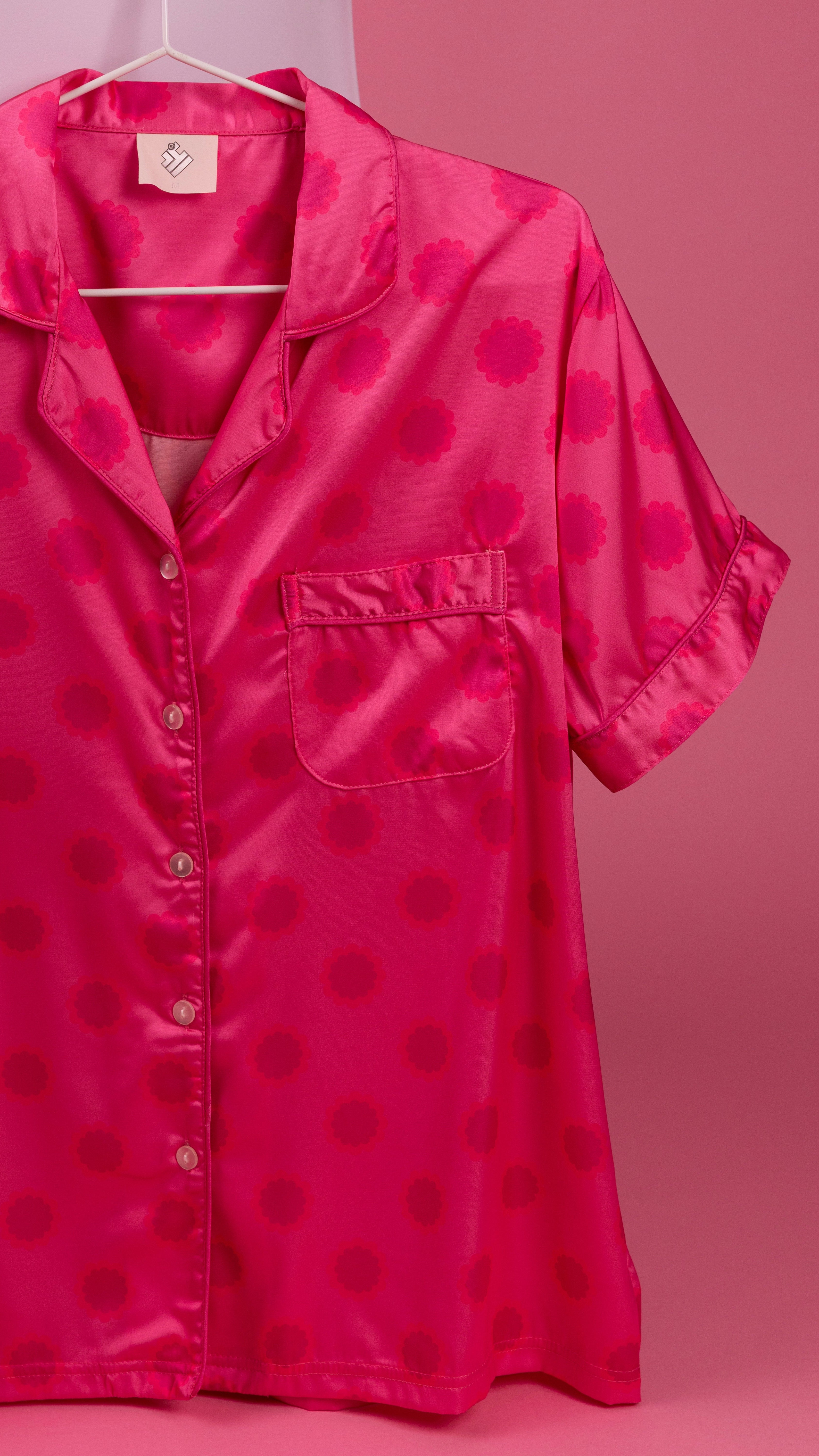 A bright pink short-sleeve button-up shirt with a darker pink circular dot pattern, hanging on a white hanger against a solid pink background.