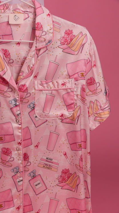 A pink short-sleeve button-up pajama shirt with an all-over print of pink handbags, perfume bottles, high heels, and flowers, shown hanging against a solid pink background.