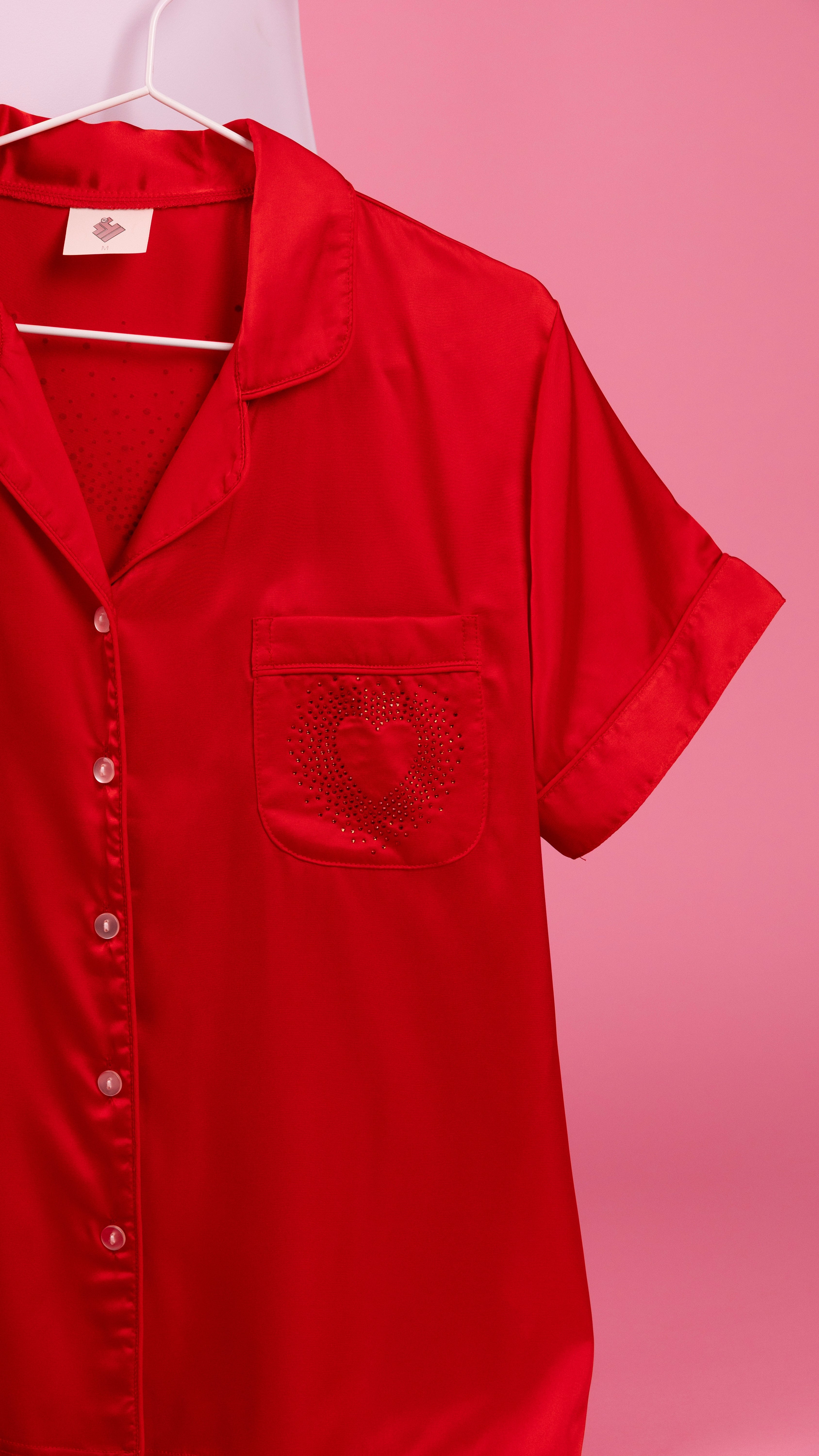 A bright red short-sleeve button-up shirt with a chest pocket, hanging on a white hanger against a pink background.