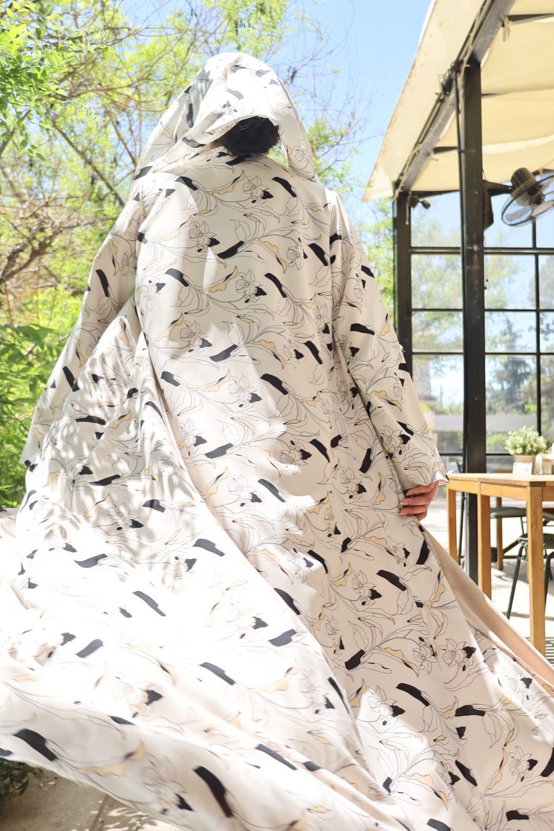 A large white fabric cover or sheet with a black abstract leaf-like pattern, being held up outdoors near a window and green trees.
