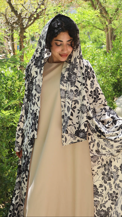 A woman wearing a beige dress with a black and white floral print outer layer, photographed outdoors in a garden setting.