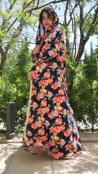 A woman wearing a long navy maxi dress with a vibrant orange, red, and yellow floral print, standing outdoors on a paved walkway with green trees in the background.