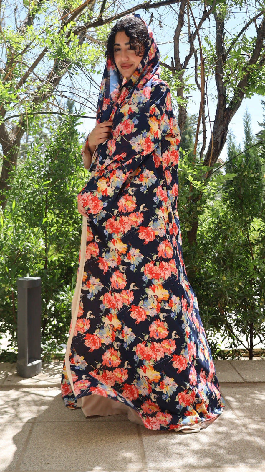 A woman wearing a long navy maxi dress with a vibrant orange, red, and yellow floral print, standing outdoors on a paved walkway with green trees in the background.