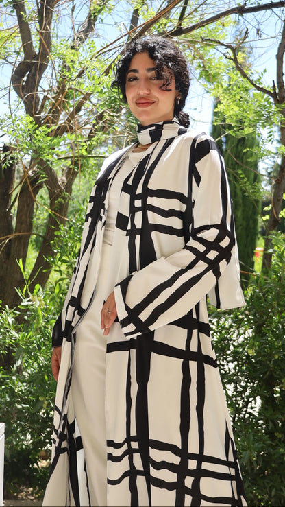 A person wearing a cream-colored long-sleeve top with a bold black geometric plaid pattern, photographed outdoors under green trees.
