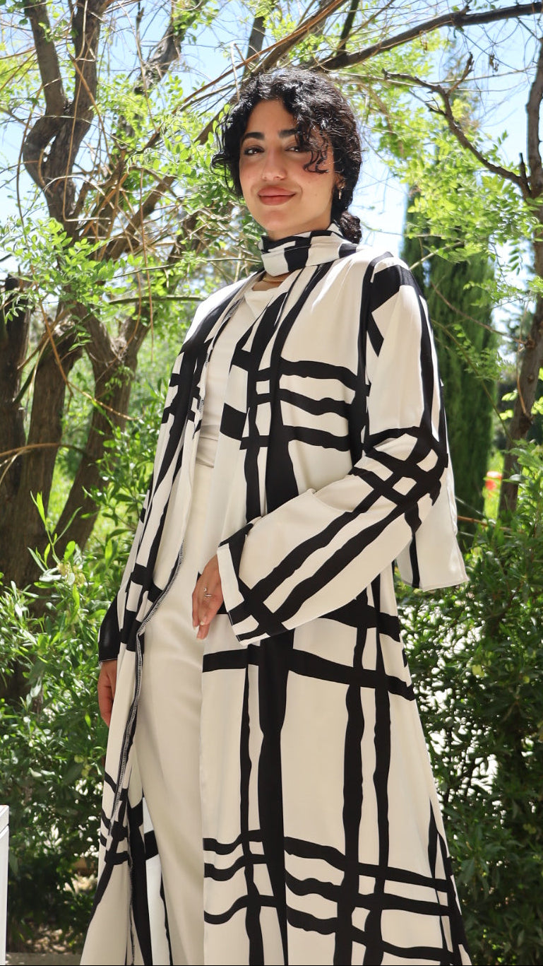 A person wearing a cream-colored long-sleeve top with a bold black geometric plaid pattern, photographed outdoors under green trees.