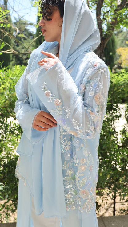 A woman wearing a light blue embroidered traditional outfit with a matching headscarf, standing outdoors in front of green trees.