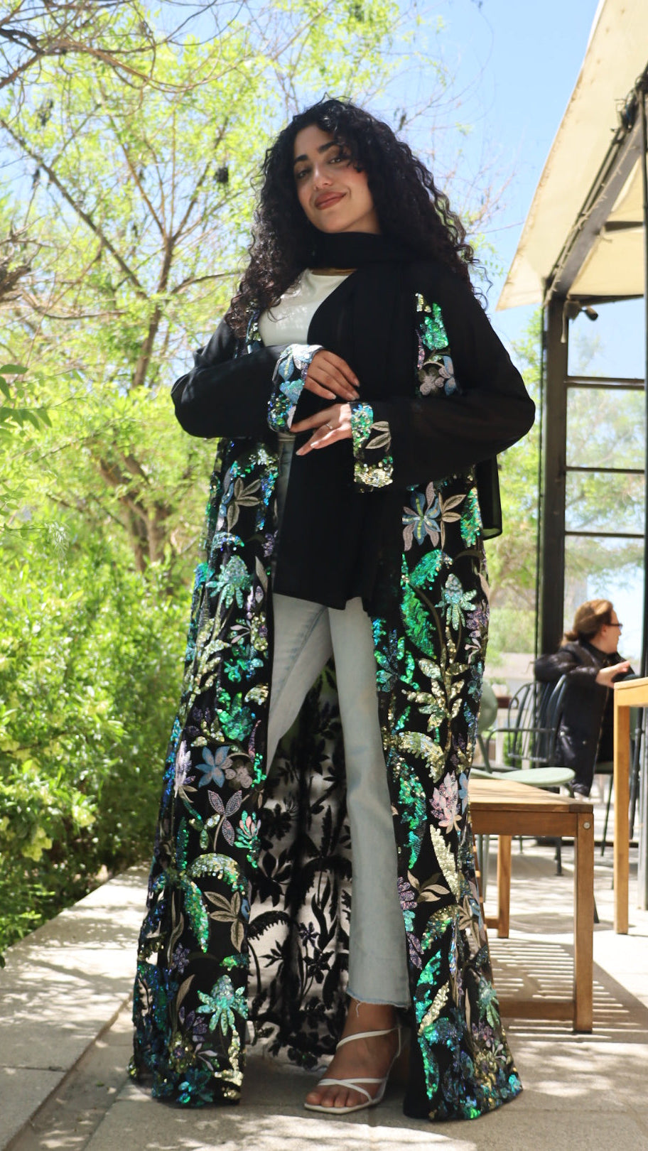 A woman wearing a long black embroidered abaya with green and white floral patterns, paired with light gray pants, standing outdoors on a patio with trees in the background.