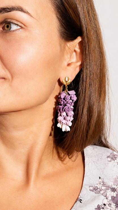 A woman with brown hair is shown in a close-up portrait wearing a light gray floral top and matching purple and white flower-shaped drop earrings.